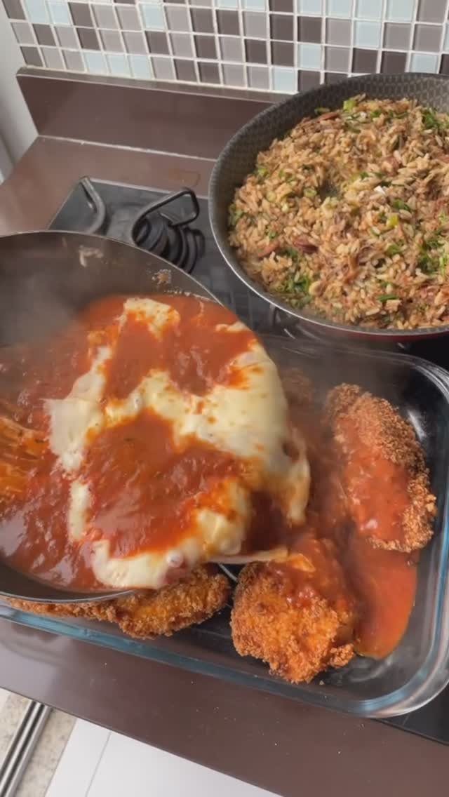Parmegiana de frango recebendo molho de tomate e queijo, com arroz na frigideira ao lado